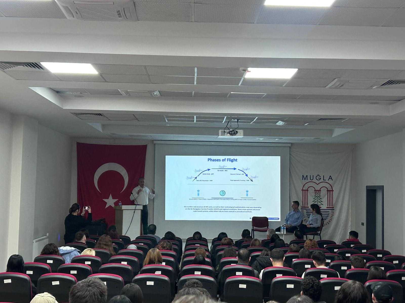 Salonun en arkasından çekilen bu geniş açılı kare, etkinliğe olan yoğun katılımı gözler önüne seriyor. Öğrencilerin dolu sıralarda pürdikkat sunumu takip etmesi ve ekrandaki "Uçuş Safhaları" şeması, seminerin akademik derinliğini ve ilgi çekiciliğini kanıtlıyor.